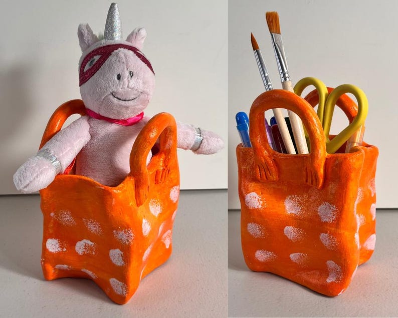 May include: Orange ceramic pencil holder with white polka dots shaped like a shopping bag. A pink unicorn plush toy is sitting inside the holder.