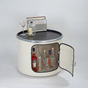 Puede incluir: Un mueble de tambor met&aacute;lico color crema con una tapa circular negra y una puerta lateral abatible. El interior contiene varias botellas de vidrio, incluida una con etiqueta de Aperol. Una radio plateada y una copa de martini est&aacute;n encima.