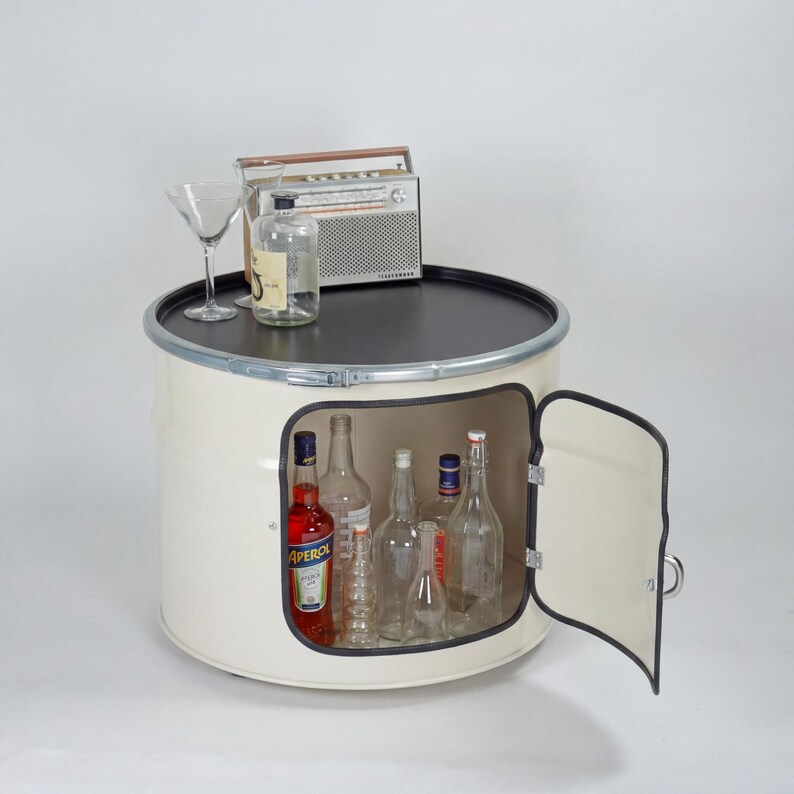 Puede incluir: Un mueble de tambor met&aacute;lico color crema con una tapa circular negra y una puerta lateral abatible. El interior contiene varias botellas de vidrio, incluida una con etiqueta de Aperol. Una radio plateada y una copa de martini est&aacute;n encima.