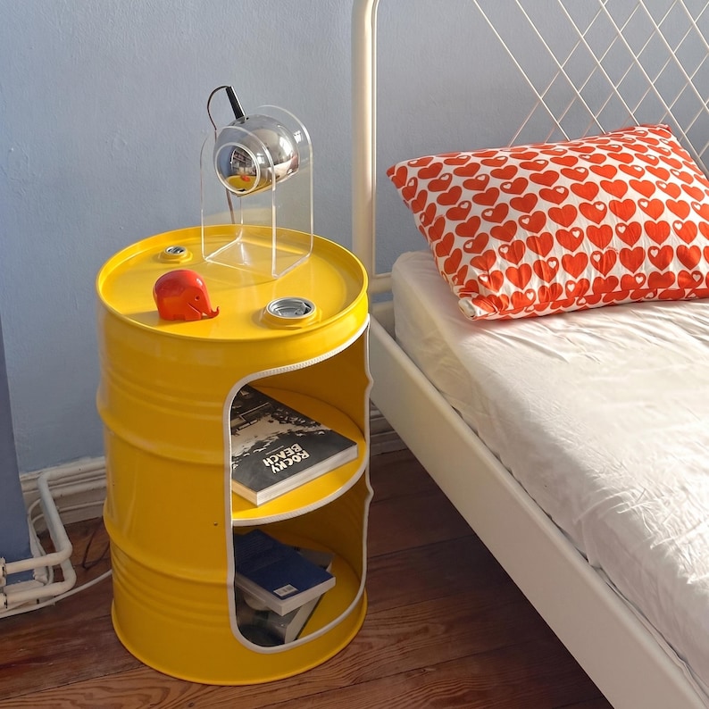 May include: A bright yellow barrel-shaped side table with two shelves. The table has a cut-out section on the front. A red decorative object and a clear lamp sit on top. Books are visible on the shelves. A bed with a heart-patterned pillow is in the background.