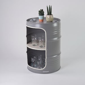 May include: A gray metal barrel repurposed as a storage unit with two shelves. The unit has a white interior and holds several clear drinking glasses. Two small potted plants sit on top. The unit is a unique piece of furniture.