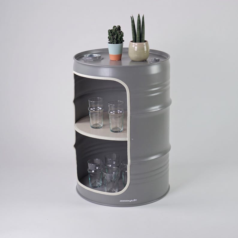 May include: A gray metal barrel repurposed as a storage unit with two shelves. The unit has a white interior and holds several clear drinking glasses. Two small potted plants sit on top. The unit is a unique piece of furniture.