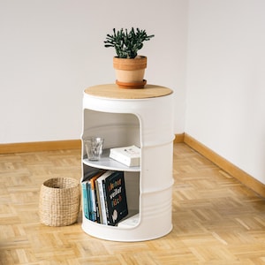May include: A white, barrel-shaped side table with a light wood top and two interior shelves. A small potted plant sits on top. The shelves hold books and a glass. A woven basket sits nearby. The table is approximately 24 inches tall.