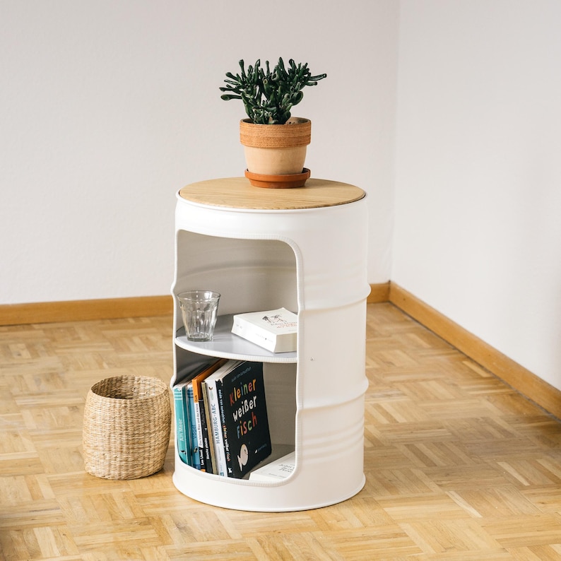 May include: A white, barrel-shaped side table with a light wood top and two interior shelves. A small potted plant sits on top. The shelves hold books and a glass. A woven basket sits nearby. The table is approximately 24 inches tall.