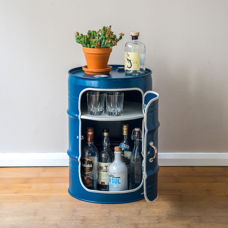 Puede incluir: Un barril de metal azul reutilizado como carrito de bar con un estante y una puerta. El carrito est&aacute; lleno de botellas de licor y vasos. Un peque&ntilde;o suculento en maceta se coloca encima del carrito.
