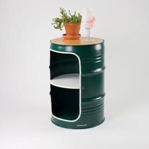 May include: A dark green metal barrel repurposed as a side table. It features a wooden top, a white interior shelf, and a cut-out design. A potted plant and a wine glass with a decorative paper are on top. The barrel is approximately 24 inches tall.