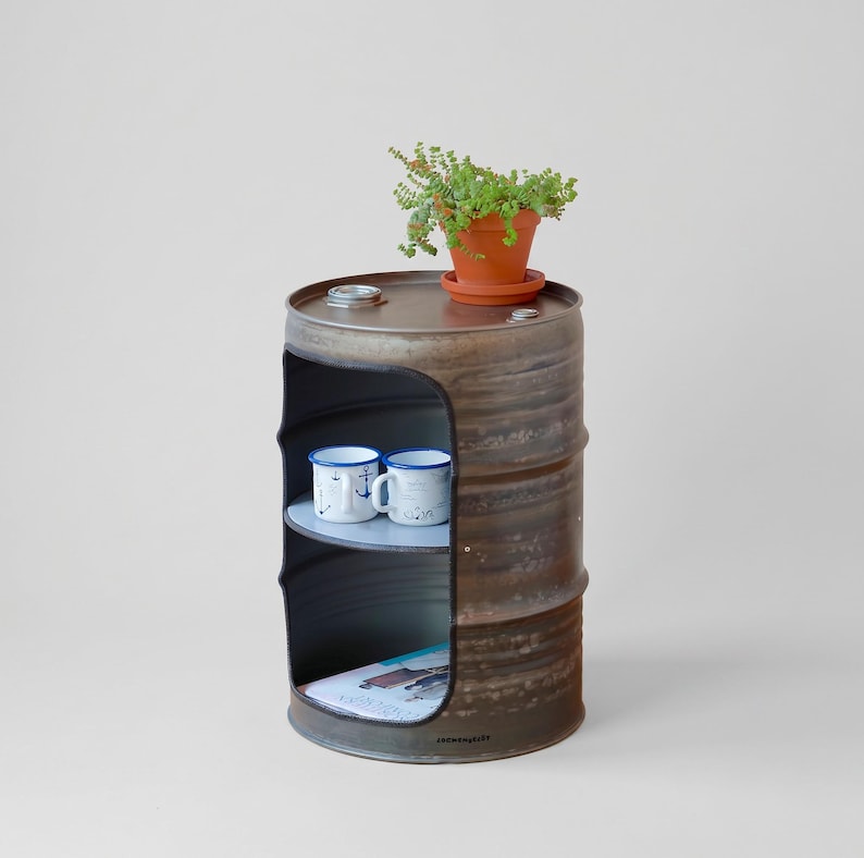 May include: A repurposed metal barrel with a cut-out section, serving as a unique side table. It features two shelves, holding two white mugs with blue nautical designs and a magazine. A small potted plant sits on top.