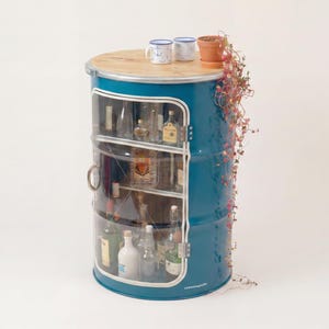 May include: A teal metal barrel cabinet with a light wood top, featuring a glass-fronted door and internal shelving holding various glass bottles. Two white mugs and a small terracotta pot with a trailing plant sit on the flat wooden surface.