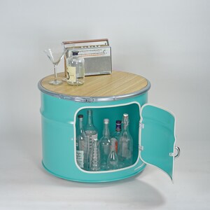 Puede incluir: Un mueble de tambor met&aacute;lico de color turquesa con una tapa circular de madera clara y una puerta abatible. El interior contiene varias botellas de vidrio transparente. Encima hay una radio de estilo vintage, una botella y una copa de martini.
