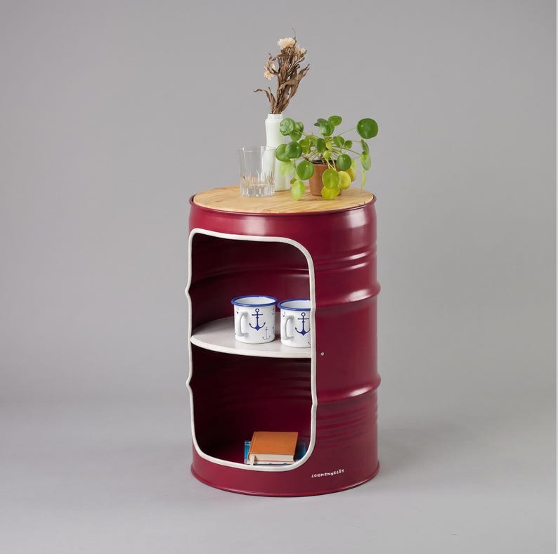 May include: A burgundy barrel-shaped side table with a wooden top and a white interior shelf. Two anchor-themed mugs sit on the shelf. The top holds a small plant, a vase with dried flowers, and a glass. The table is a unique home decor item.