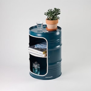 May include: A teal blue barrel-shaped side table with a white trim and a cut-out shelf. The top holds a small potted plant, a glass of water, and a small coaster. The shelf contains books and two glasses. The text "LOCHENGELLDT" is printed on the side.