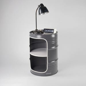 May include: A gray metal barrel-shaped side table with a white interior shelf and a white trim. A black desk lamp and a digital clock sit on top. The lamp has a black shade and a flexible neck. The clock displays the time.