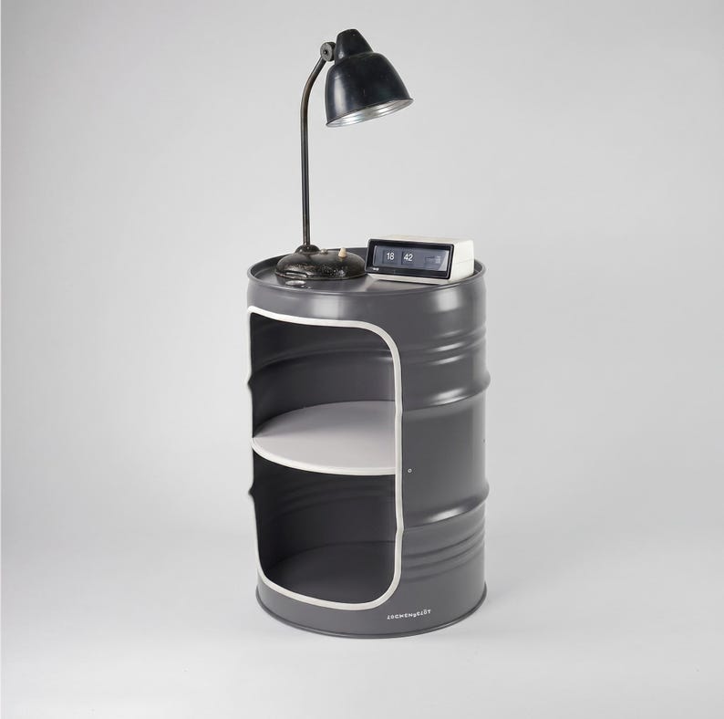 May include: A gray metal barrel-shaped side table with a white interior shelf and a white trim. A black desk lamp and a digital clock sit on top. The lamp has a black shade and a flexible neck. The clock displays the time.