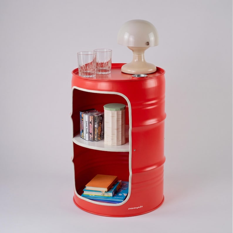 May include: A red barrel-shaped side table with a white interior shelf. The top holds two clear glasses and a beige mushroom-shaped lamp. The shelf displays CDs, a decorative container, and books. The bottom of the barrel has the text "cosmopolitan".