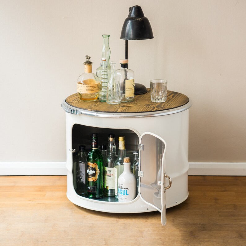 Puede incluir: Un mueble cil&iacute;ndrico de metal blanco con una tapa de madera oscura, que cuenta con una puerta abatible y un estante interior. La unidad contiene varias botellas de vidrio y est&aacute; coronada por una l&aacute;mpara de escritorio negra y botellas vac&iacute;as.