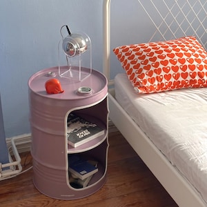 May include: A lilac-colored barrel-shaped nightstand with two shelves and a circular top. The top holds a small chrome lamp and a red elephant figurine. A pillow with a red heart pattern rests on a white bed.