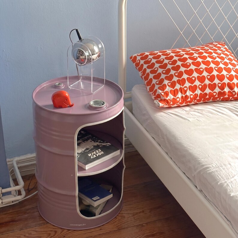 May include: A lilac-colored barrel-shaped nightstand with two shelves and a circular top. The top holds a small chrome lamp and a red elephant figurine. A pillow with a red heart pattern rests on a white bed.