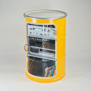 Peut inclure: Un fût métallique cylindrique jaune vif reconverti en meuble de rangement. Il est doté d'une porte en acrylique transparent avec une poignée argentée, révélant des étagères internes contenant du matériel audio vintage et des livres.