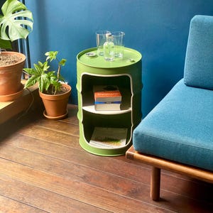 May include: A green, upcycled barrel side table with white trim and two interior shelves. The tabletop holds two clear glasses and a small bottle. Books and a magazine are on the shelves. The table is next to a blue couch.