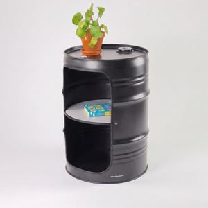 May include: A black metal barrel repurposed as a side table. The table features a cut-out section with a shelf and a potted plant on top. A book rests on the shelf. The table has a industrial aesthetic.
