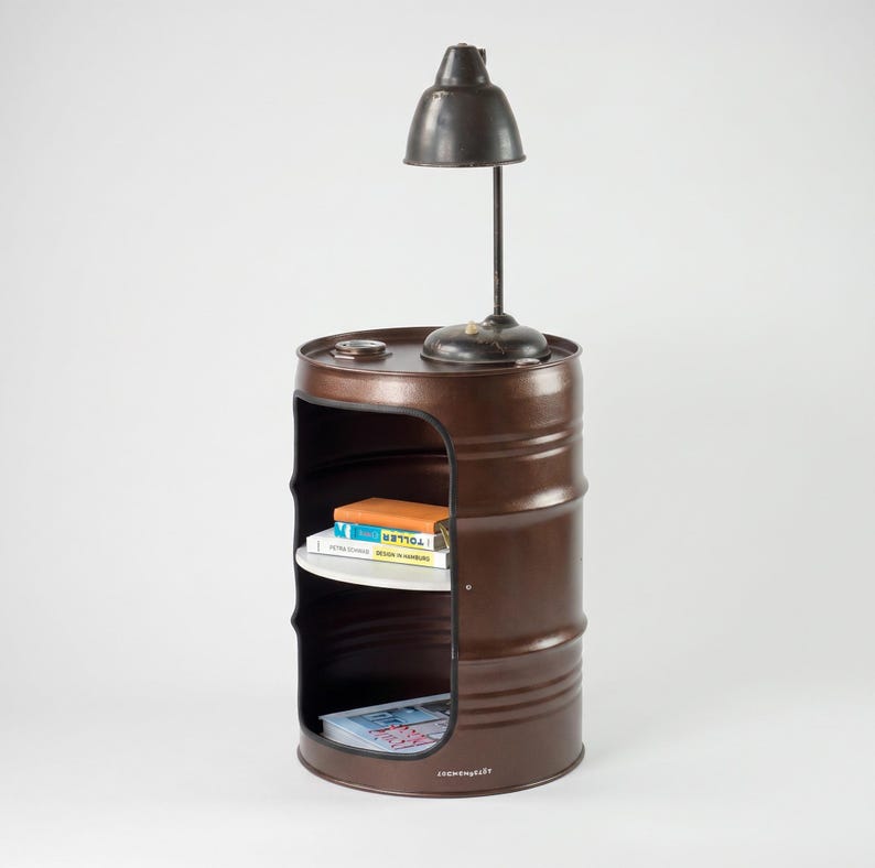 May include: A brown metal barrel repurposed as a side table with a built-in shelf and a small lamp on top. The barrel has a cut-out section for storage, and the lamp has a dark metal shade. Books are visible on the shelf.