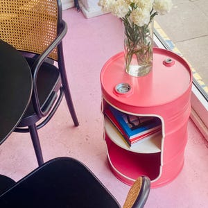 May include: A pink metal barrel side table with a white trim and a shelf. The table has a round top with a clear glass vase of white flowers. Books are visible on the shelf. The table is in a room with a pink floor.