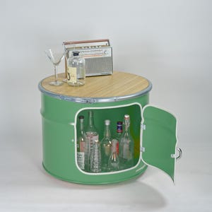Puede incluir: Un mueble de tambor met&aacute;lico verde con tapa redonda de madera, que cuenta con una puerta abatible y un espacio de almacenamiento interior. Encima hay una radio de estilo vintage, una botella de vidrio transparente y una copa de martini.
