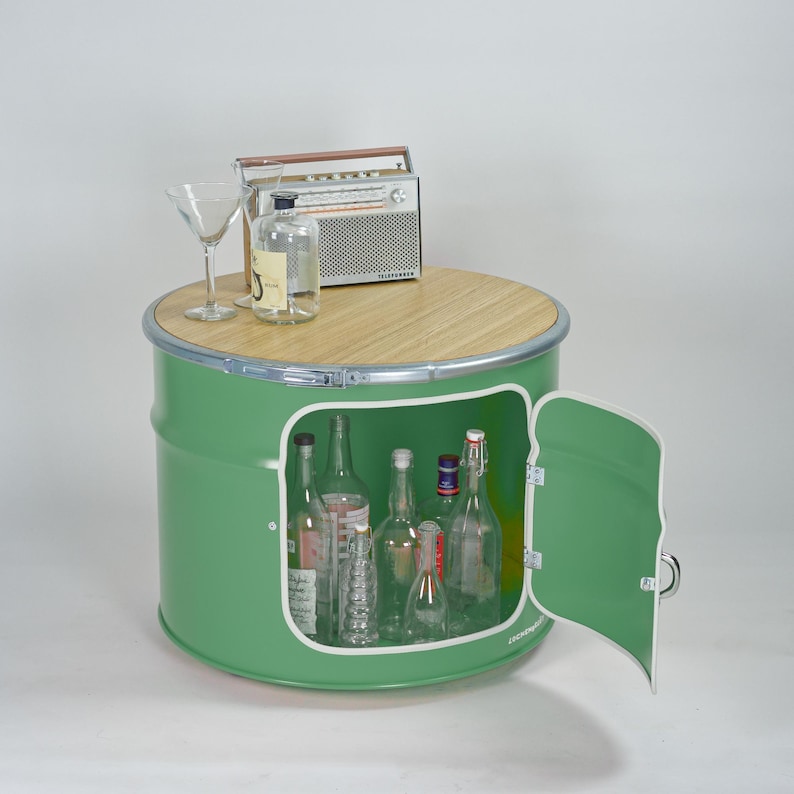 Puede incluir: Un mueble de tambor met&aacute;lico verde con tapa redonda de madera, que cuenta con una puerta abatible y un espacio de almacenamiento interior. Encima hay una radio de estilo vintage, una botella de vidrio transparente y una copa de martini.
