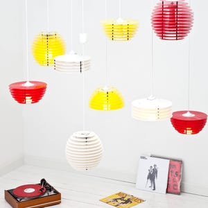 Puede incluir: Una colección de modernas lámparas colgantes en rojo, amarillo y blanco, suspendidas del techo. Las luces tienen un diseño circular en capas. Un tocadiscos vintage y discos de vinilo están en primer plano, lo que sugiere un tema retro.