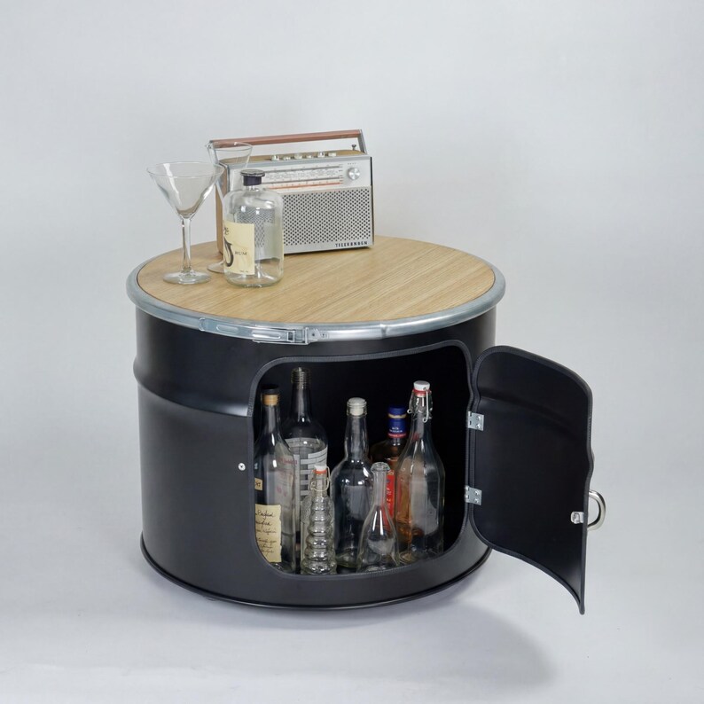 Puede incluir: Un mueble cil&iacute;ndrico de metal negro con tapa de madera clara, que cuenta con una puerta abatible que revela un espacio de almacenamiento con varias botellas de vidrio. Sobre la superficie superior hay una radio vintage y una copa de martini.