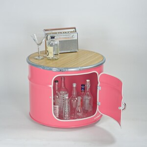Puede incluir: Un mueble de metal rosa con forma de barril, tapa circular de madera y puerta abatible que revela un espacio de almacenamiento. Encima hay una radio de estilo vintage, una botella de vidrio transparente y una copa de martini. Dentro hay botellas vac&iacute;as.
