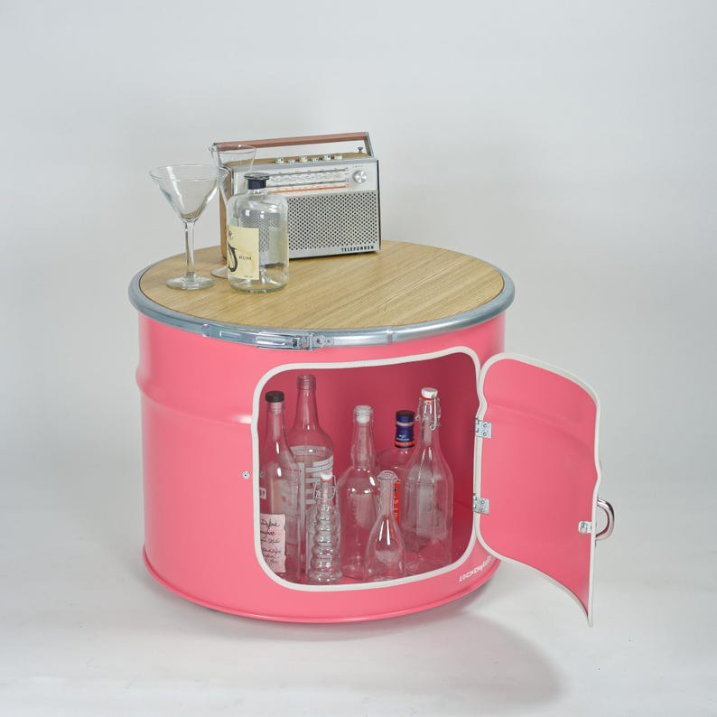 Puede incluir: Un mueble de metal rosa con forma de barril, tapa circular de madera y puerta abatible que revela un espacio de almacenamiento. Encima hay una radio de estilo vintage, una botella de vidrio transparente y una copa de martini. Dentro hay botellas vac&iacute;as.