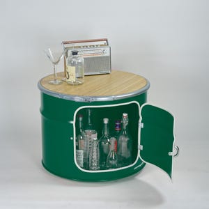 Puede incluir: Un mueble de almacenamiento cil&iacute;ndrico de metal verde con una tapa circular de madera clara, que cuenta con una puerta abatible con borde blanco. Dentro hay varias botellas de vidrio. Encima hay una radio vintage, una botella y una copa.