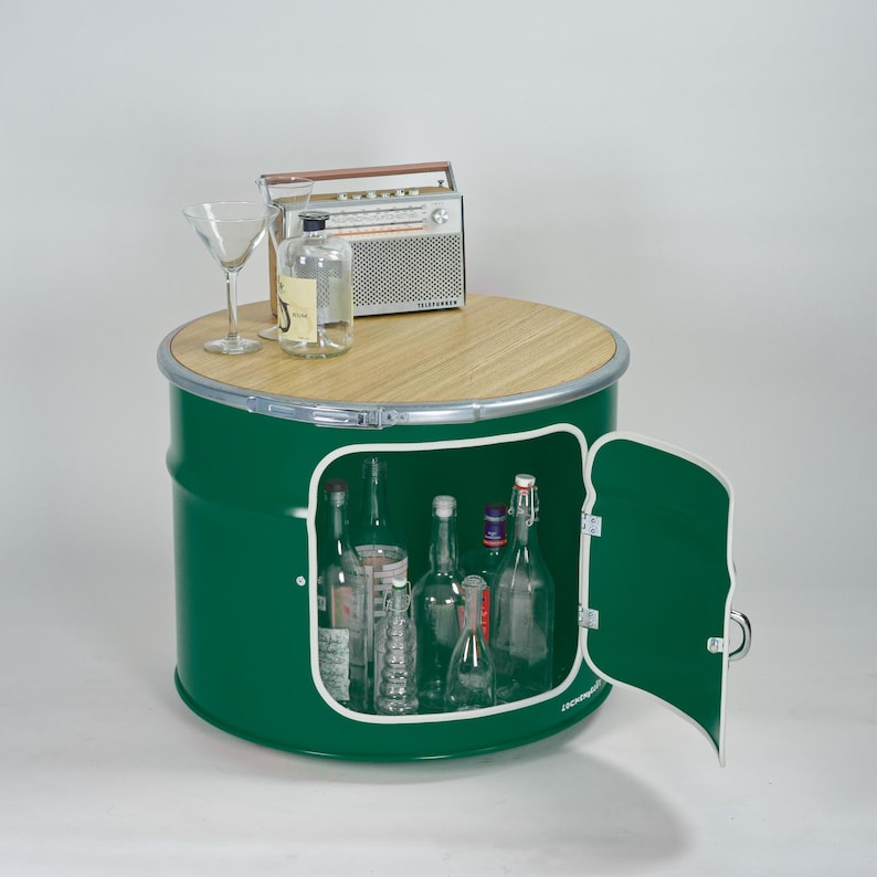 Puede incluir: Un mueble de almacenamiento cil&iacute;ndrico de metal verde con una tapa circular de madera clara, que cuenta con una puerta abatible con borde blanco. Dentro hay varias botellas de vidrio. Encima hay una radio vintage, una botella y una copa.