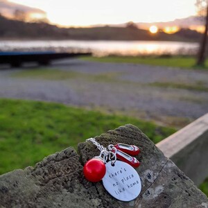 Puede incluir: Collar de plata con una cuenta roja, un dije de zapatos rojos y un dije de plata con el texto "there's no place like home".