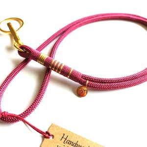 Könnte beinhalten: Ein pinkfarbenes Hundehalsband mit goldfarbener Metallhardware. Das Halsband hat einen goldfarbenen Verschluss und einen kleinen Anhänger. Das Seil ist mit goldfarbenem Draht umwickelt. Ein kleines Etikett ist am Halsband befestigt.
