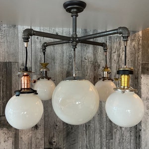 Puede incluir: Lámpara de techo de estilo industrial con cinco pantallas de globo blancas. La lámpara presenta un marco de metal oscuro con accesorios de tubería y detalles en cobre y latón. Las pantallas están espaciadas uniformemente.
