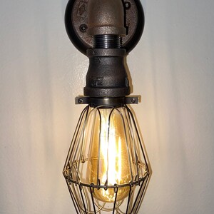 May include: Industrial-style wall sconce featuring a black metal pipe design. The fixture includes a cage-like shade surrounding a warm, glowing Edison bulb. The sconce is mounted on a circular black base.