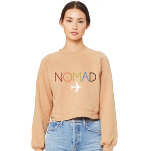 Könnte beinhalten: Ein beiger, kurzer Pullover mit dem Wort "NOMAD" in bunten Buchstaben und einem weißen Flugzeug-Motiv darunter.