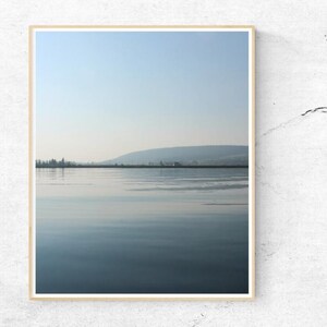 May include: A framed print depicting a tranquil lake scene under a pale blue sky. The water mirrors the sky and distant hills, producing a calming, natural aesthetic. The print is enclosed in a simple, light-coloured wooden frame.