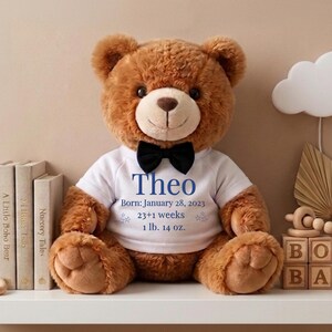 May include: A brown teddy bear wearing a white t-shirt with the name "Theo" and birth details. The bear has a black bow tie and is sitting on a white shelf with baby-themed decor, including wooden blocks that spell "BOHO BABY".