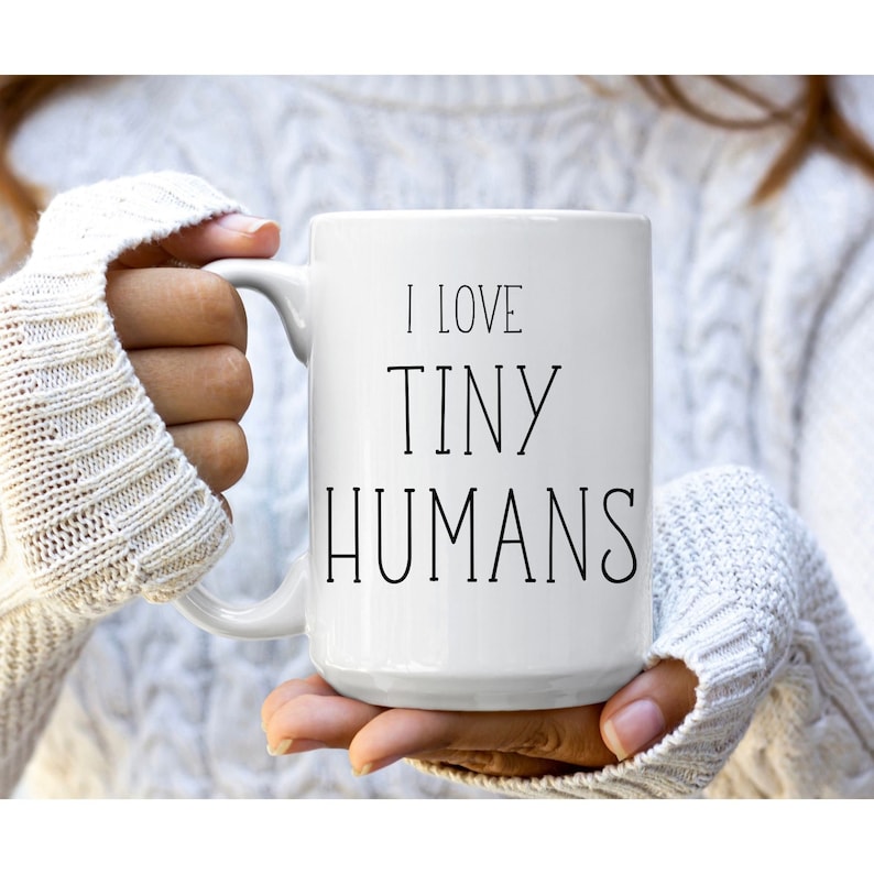 Może przedstawiać: Biały ceramiczny kubek z czarnym napisem "I LOVE TINY HUMANS". Kubek ma duży uchwyt i jest trzymany przez osobę w białym swetrze. Kubek jest przeznaczony do gorących napoj&oacute;w.