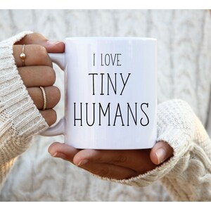 Może przedstawiać: Biały ceramiczny kubek z czarnym napisem "I LOVE TINY HUMANS". Kubek jest trzymany przez osobę ubraną w biały sweter. Kubek ma uchwyt po prawej stronie.
