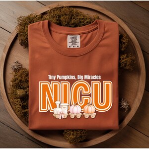 May include: A burnt orange t-shirt featuring the text "Tiny Pumpkins, Big Miracles" above the word "NICU" in large white and orange letters. Below the text is a graphic of a toy train carrying two pumpkins, one light pink and one orange.