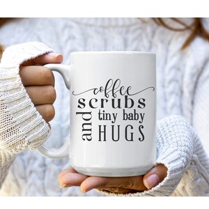 May include: White ceramic coffee mug with black text that reads "coffee scrubs and tiny baby hugs".