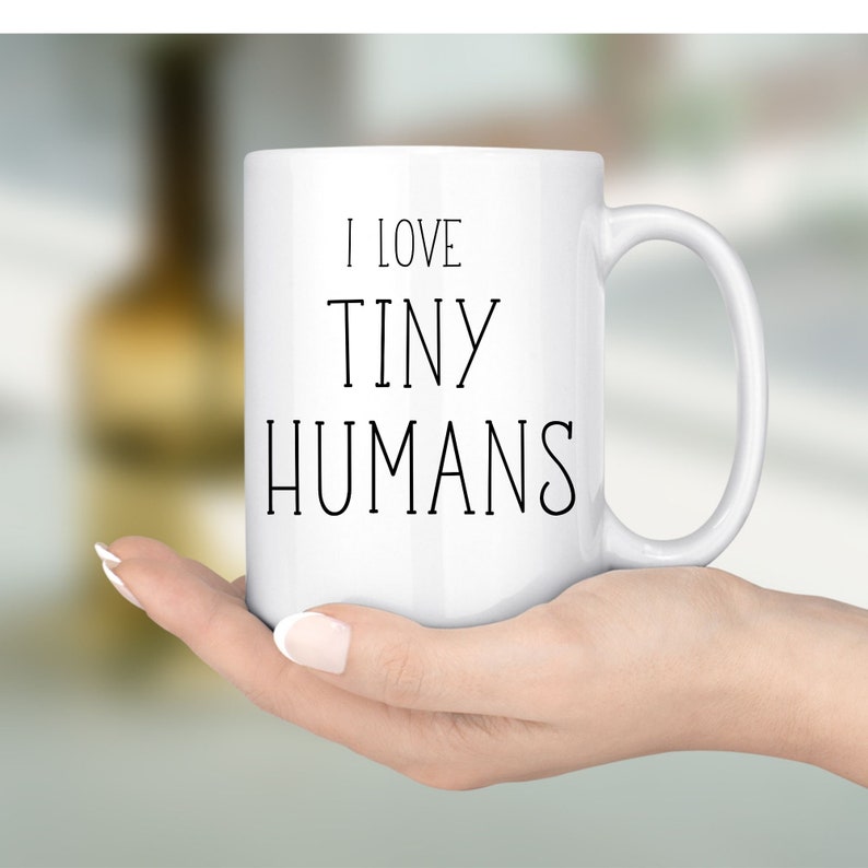 Może przedstawiać: Biały ceramiczny kubek z czarnym napisem "I LOVE TINY HUMANS". Kubek ma zakrzywiony uchwyt i jest trzymany przez osobę. Tło jest rozmyte, co sugeruje miękkie ustawienie ostrości.