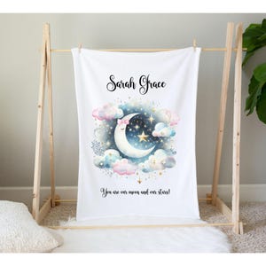 Puede incluir: Una manta de beb&eacute; blanca con un nombre personalizado "Sarah Grace" y una ilustraci&oacute;n de acuarela de una luna creciente con un lazo rosa, rodeada de nubes y estrellas. El texto "You are our moon and our stars!" est&aacute; impreso debajo de la ilustraci&oacute;n.