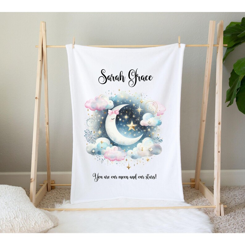 Puede incluir: Una manta de beb&eacute; blanca con un nombre personalizado "Sarah Grace" y una ilustraci&oacute;n de acuarela de una luna creciente con un lazo rosa, rodeada de nubes y estrellas. El texto "You are our moon and our stars!" est&aacute; impreso debajo de la ilustraci&oacute;n.