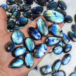 May include: A collection of blue and green labradorite gemstone cabochons, with iridescent flashes of color. The cabochons are oval shaped and have a smooth, polished finish.  The cabochons are arranged in a hand, with the text "Nikhat Gems" visible in the background.