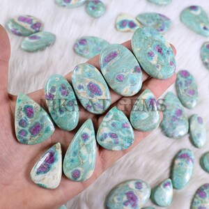 May include: A collection of polished gemstones in various shapes, including ovals and teardrops. The stones feature a teal base color with vibrant magenta and cream-colored inclusions. The gemstones are displayed on a white, textured surface.